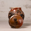 A handmade ceramic jar with a matching lid, featuring a glossy brown and amber glaze with black and terracotta mottled patterns, set against a textured light gray background.
