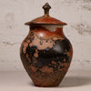 A handmade ceramic lidded jar with a rounded body, featuring a glossy glaze with a mottled pattern of black, terracotta orange, and brown, set against a textured light gray background.