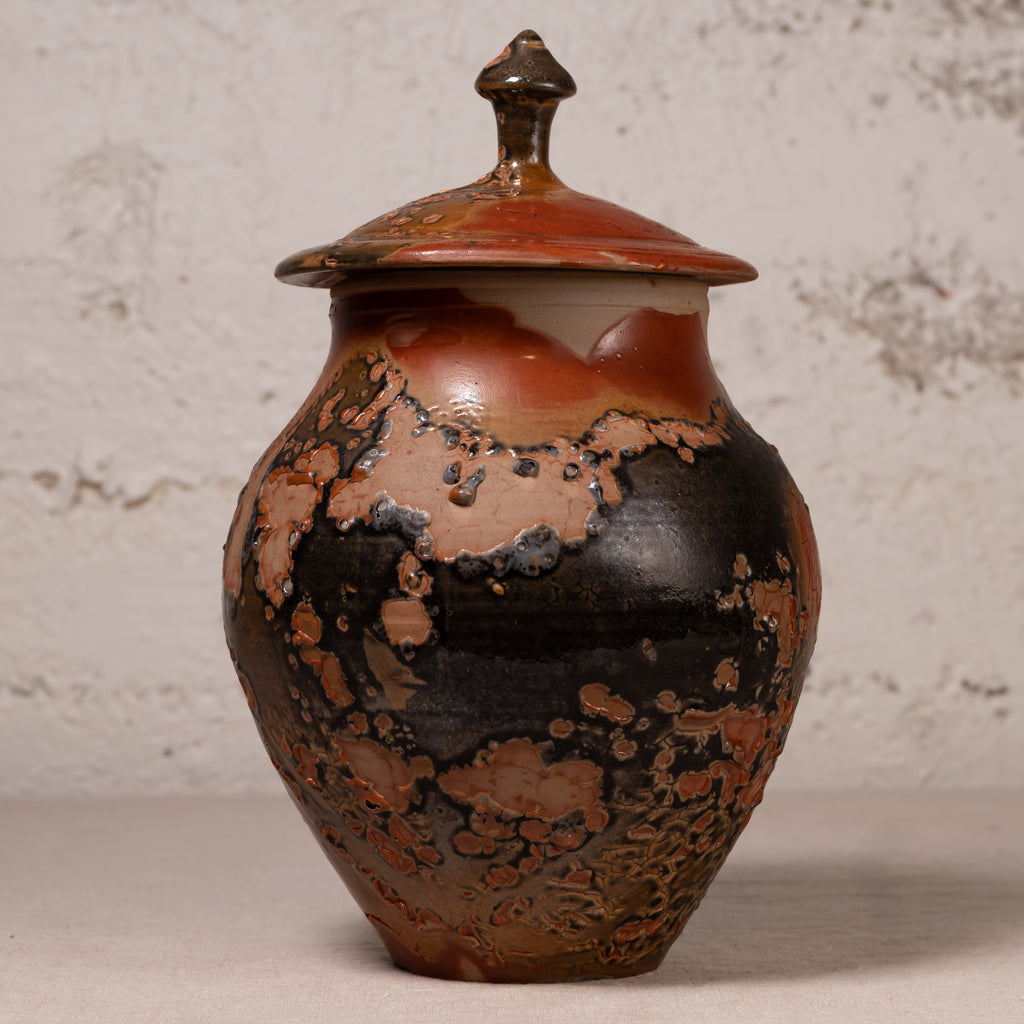 A handmade ceramic lidded jar with a rounded body, featuring a glossy glaze with a mottled pattern of black, terracotta orange, and brown, set against a textured light gray background.