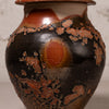 A close-up of a ceramic vase with a glossy black glaze and terracotta orange patches, set against a neutral light gray background.