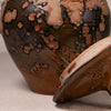 Close-up of a ceramic lidded container with a glossy brown and black glaze featuring irregular coppery orange patches, with the lid leaning against the base on a neutral fabric surface.