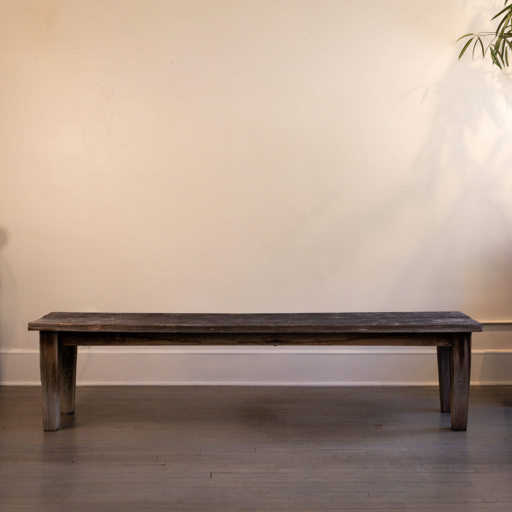 Wooden bench against a plain wall with a plant in the corner