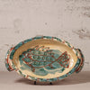 An oval ceramic serving platter with side handles, featuring a hand-painted fish design in teal and red on a beige background, set against a textured off-white wall.