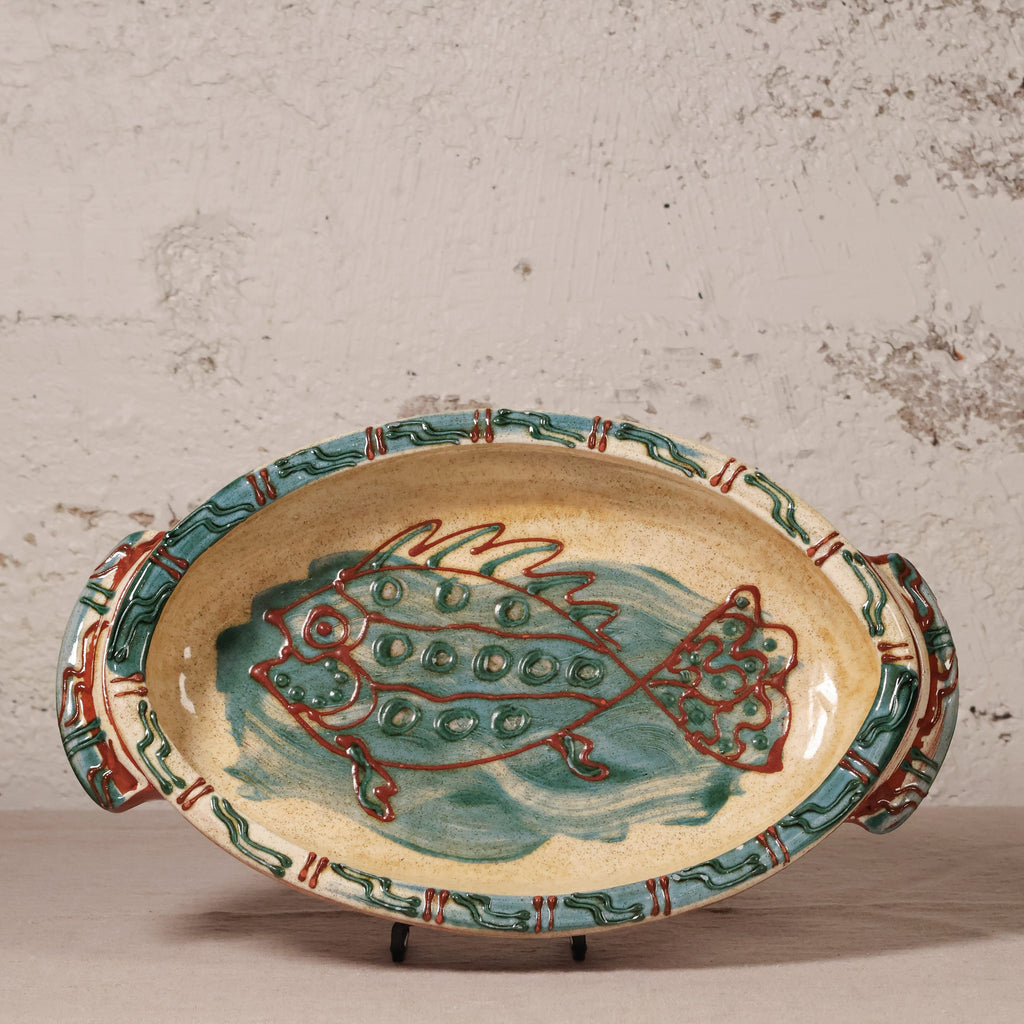 An oval ceramic serving platter with side handles, featuring a hand-painted fish design in teal and red on a beige background, set against a textured off-white wall.