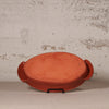 An oval terracotta clay baking dish with side handles, displayed on a small black stand against a textured, distressed off-white wall.