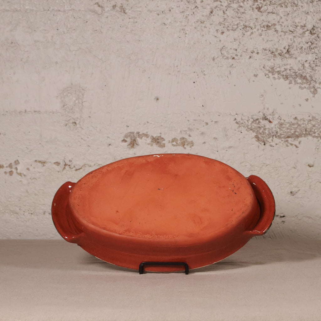An oval terracotta clay baking dish with side handles, displayed on a small black stand against a textured, distressed off-white wall.