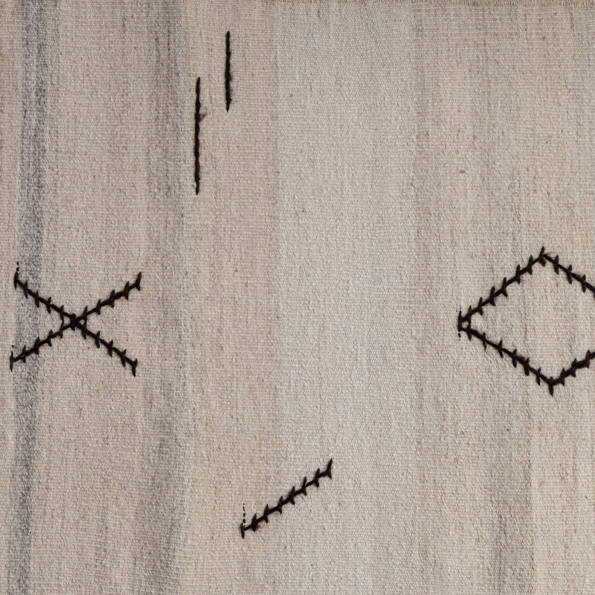 A close-up of a woven textile or rug in a warm off-white/cream tone with irregular black stitched geometric motifs, including dashes, an X, and a diamond.