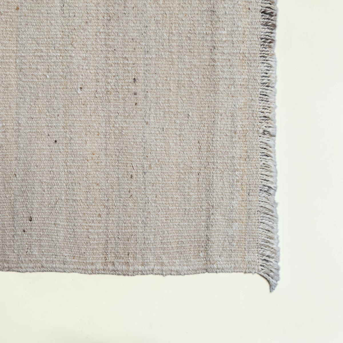 Close-up of a woven rug corner showing a textured natural fiber surface with a short fringe edge on a light neutral background.