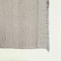 Close-up of a woven rug corner showing a textured natural fiber surface with a short fringe edge on a light neutral background.
