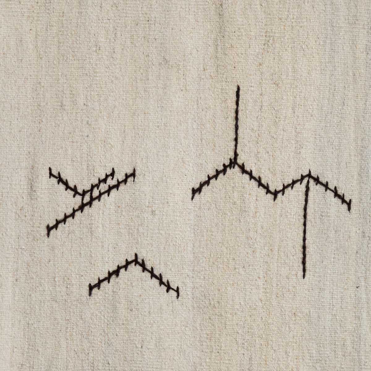 Close-up of a beige woven textile with three black embroidered geometric line motifs stitched on it.