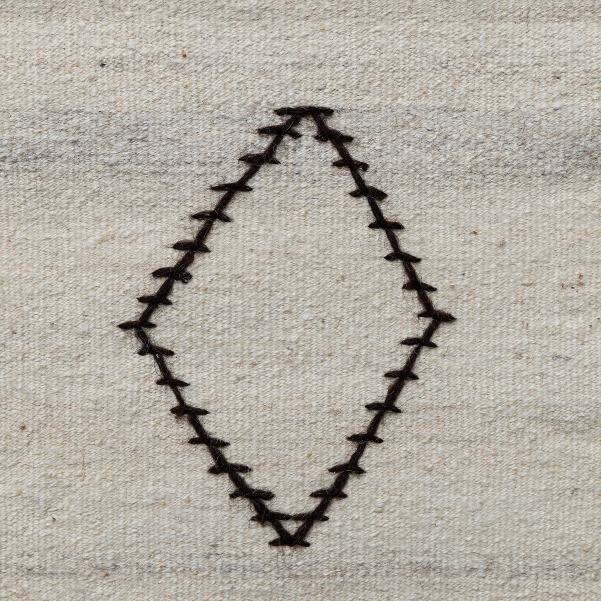 A close-up of a woven fabric surface in a light beige or off-white tone, featuring a stitched diamond-shaped outline outlined in dark brown or black thread.