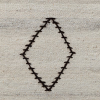 A close-up of a woven fabric surface in a light beige or off-white tone, featuring a stitched diamond-shaped outline outlined in dark brown or black thread.