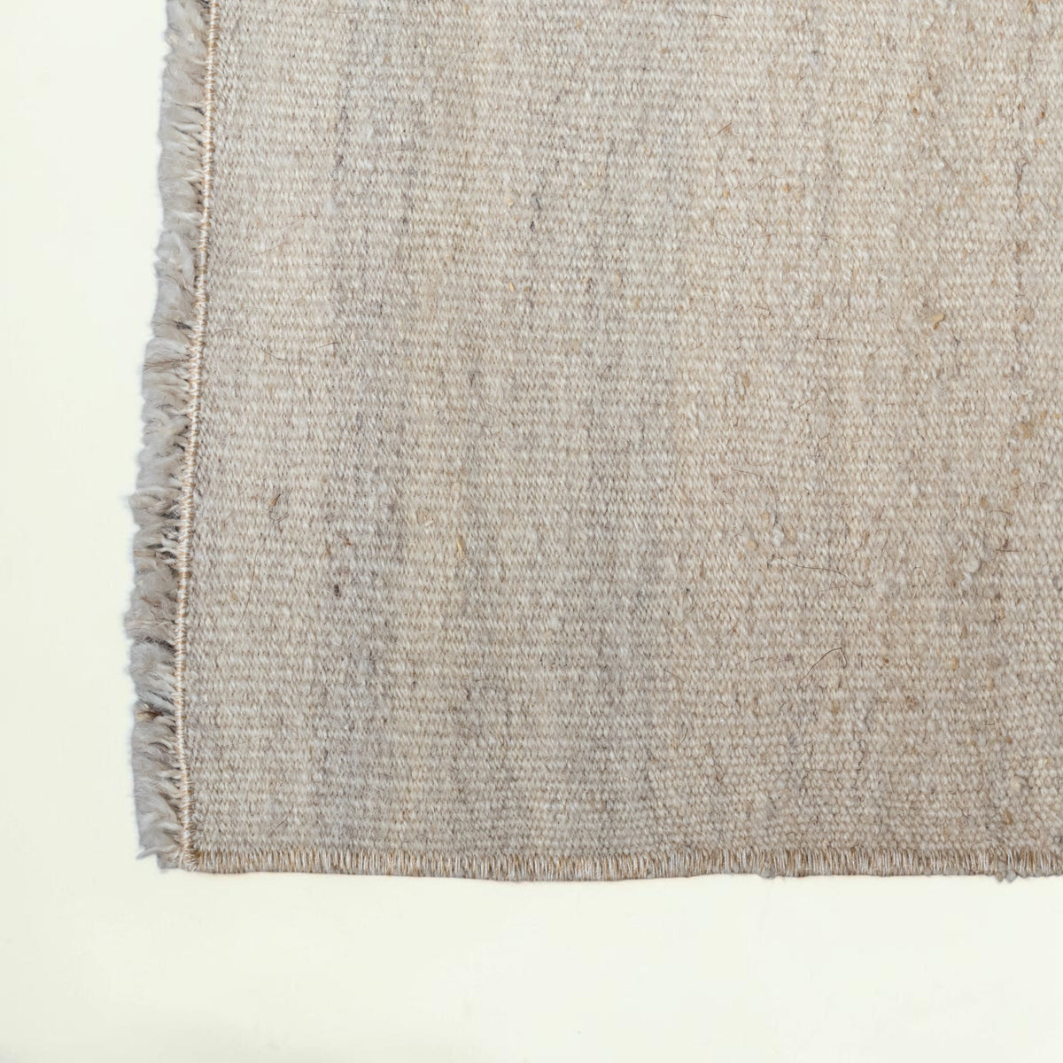 Close-up of a woven rug or textile corner showing a textured beige and light gray pattern with fringed edges on a plain white background.