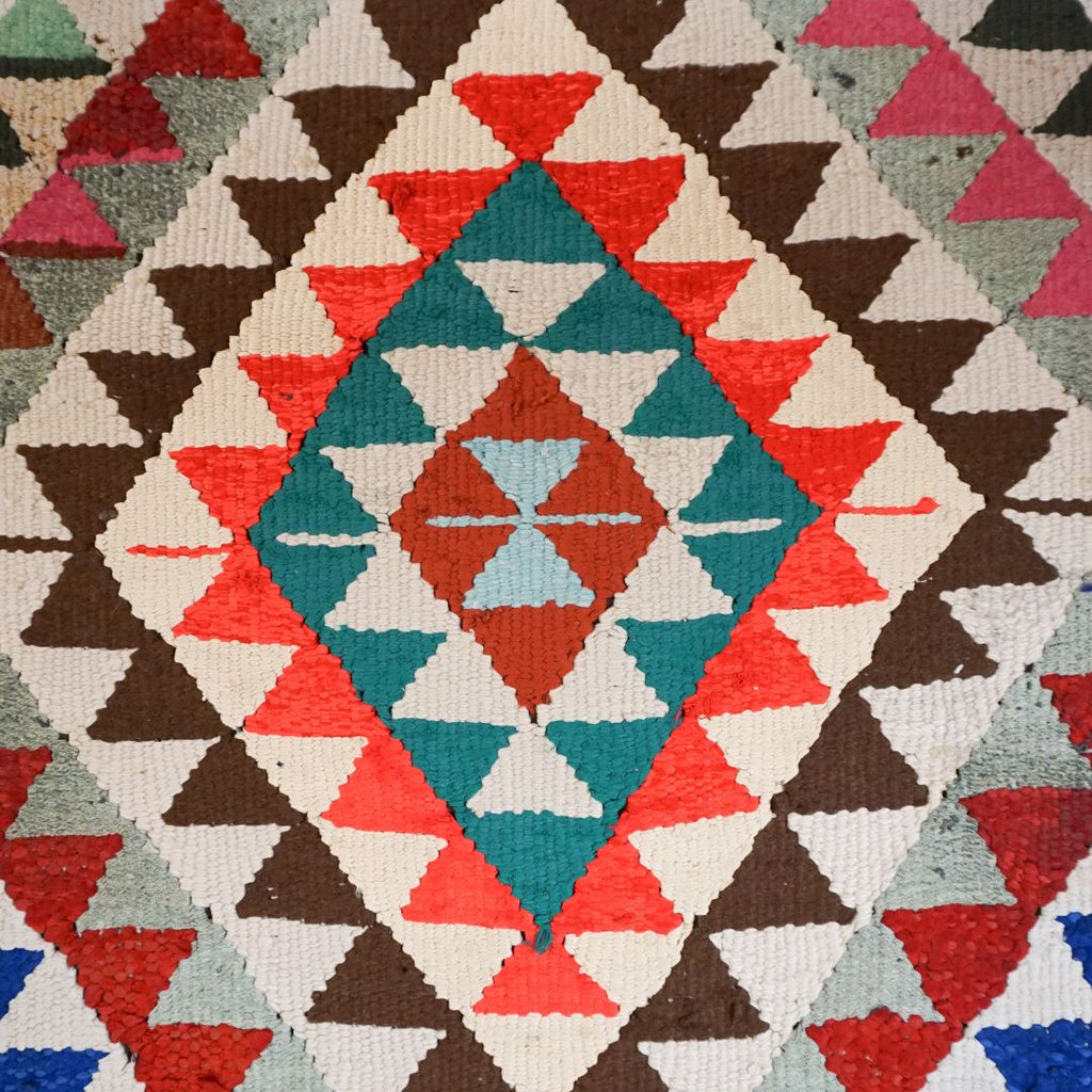 A close-up of a woven textile with a geometric diamond pattern in teal, rust red, coral, brown, cream, and light blue.