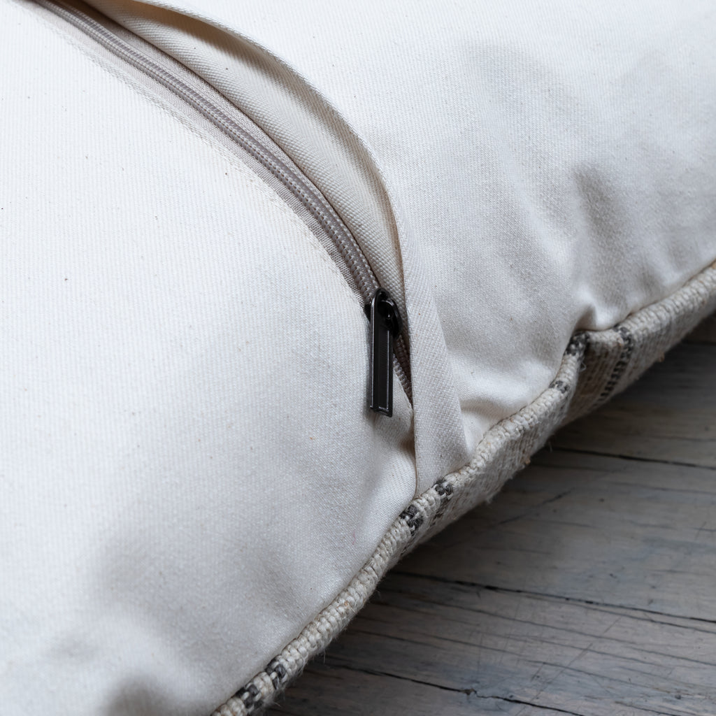 Close-up of a cream-colored fabric zippered cushion or pillow with a black zipper pull, resting on a rustic wooden surface.