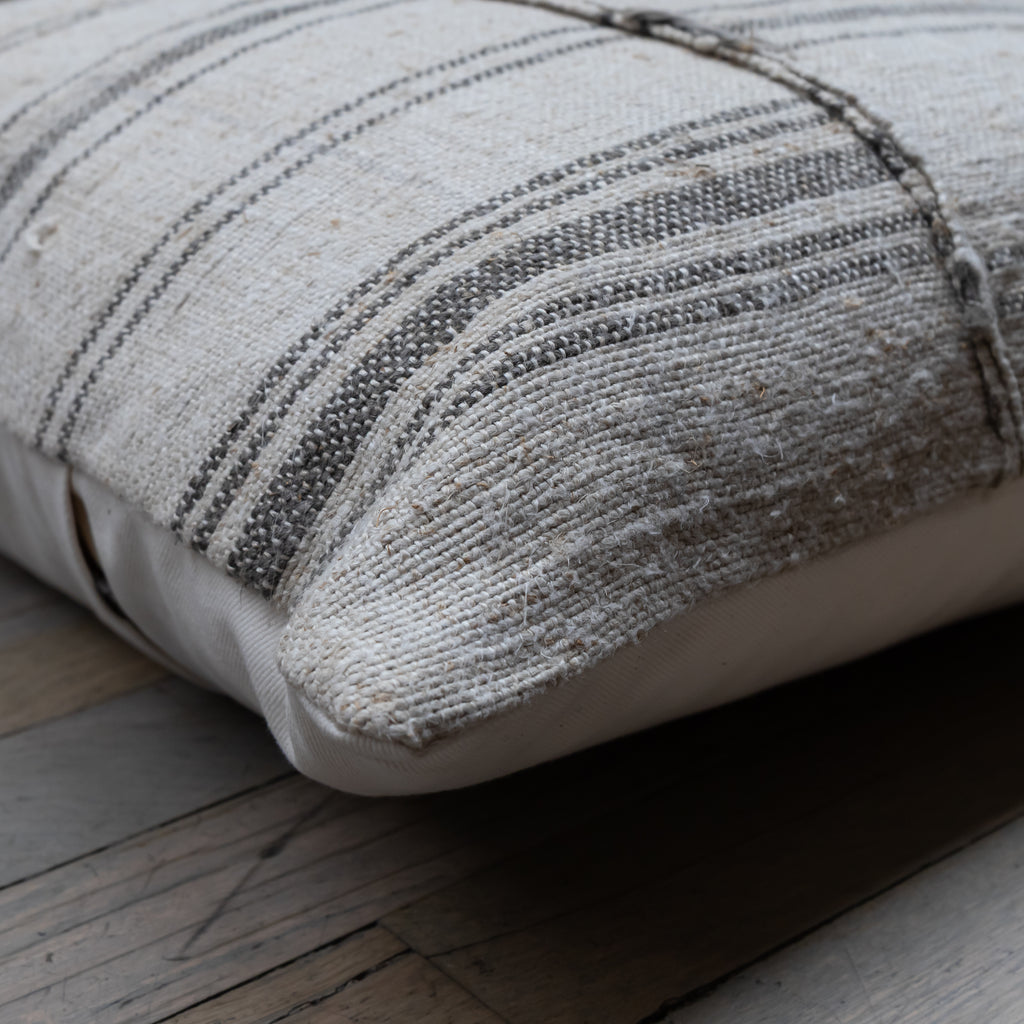 A close-up of a square throw pillow with a textured woven fabric in beige and gray stripes, resting on a rustic wooden floor.
