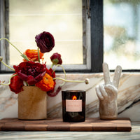 Candle with a decorative label next to flowers and a hand-shaped object on a wooden surface.