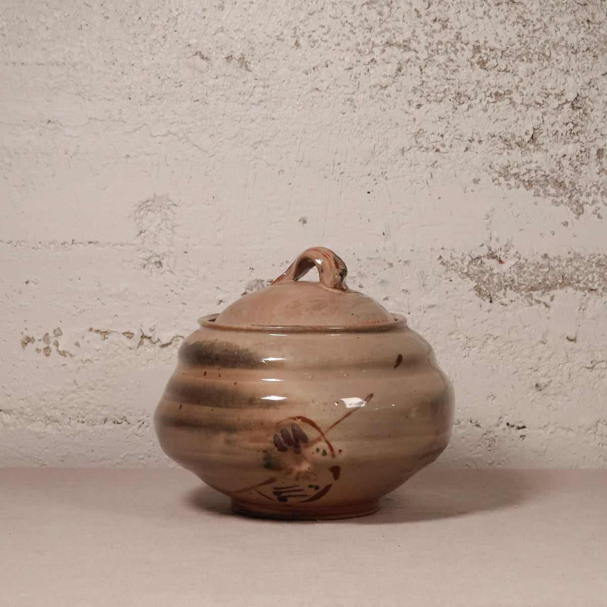 A handmade ceramic lidded jar with a rounded body, small loop handle on top, and a painted bird motif near the base, shown against a textured off-white wall.