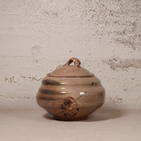 A handmade ceramic lidded jar with a rounded body, small loop handle on top, and a painted bird motif near the base, shown against a textured off-white wall.