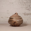 A handmade ceramic lidded jar with a rounded body, small loop handle on top, and a painted bird motif near the base, shown against a textured off-white wall.