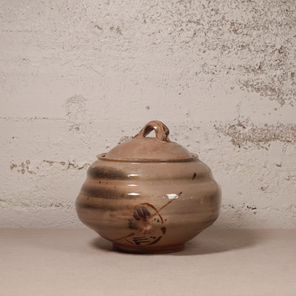 A handmade ceramic lidded jar with a rounded body, small loop handle on top, and a painted bird motif near the base, shown against a textured off-white wall.