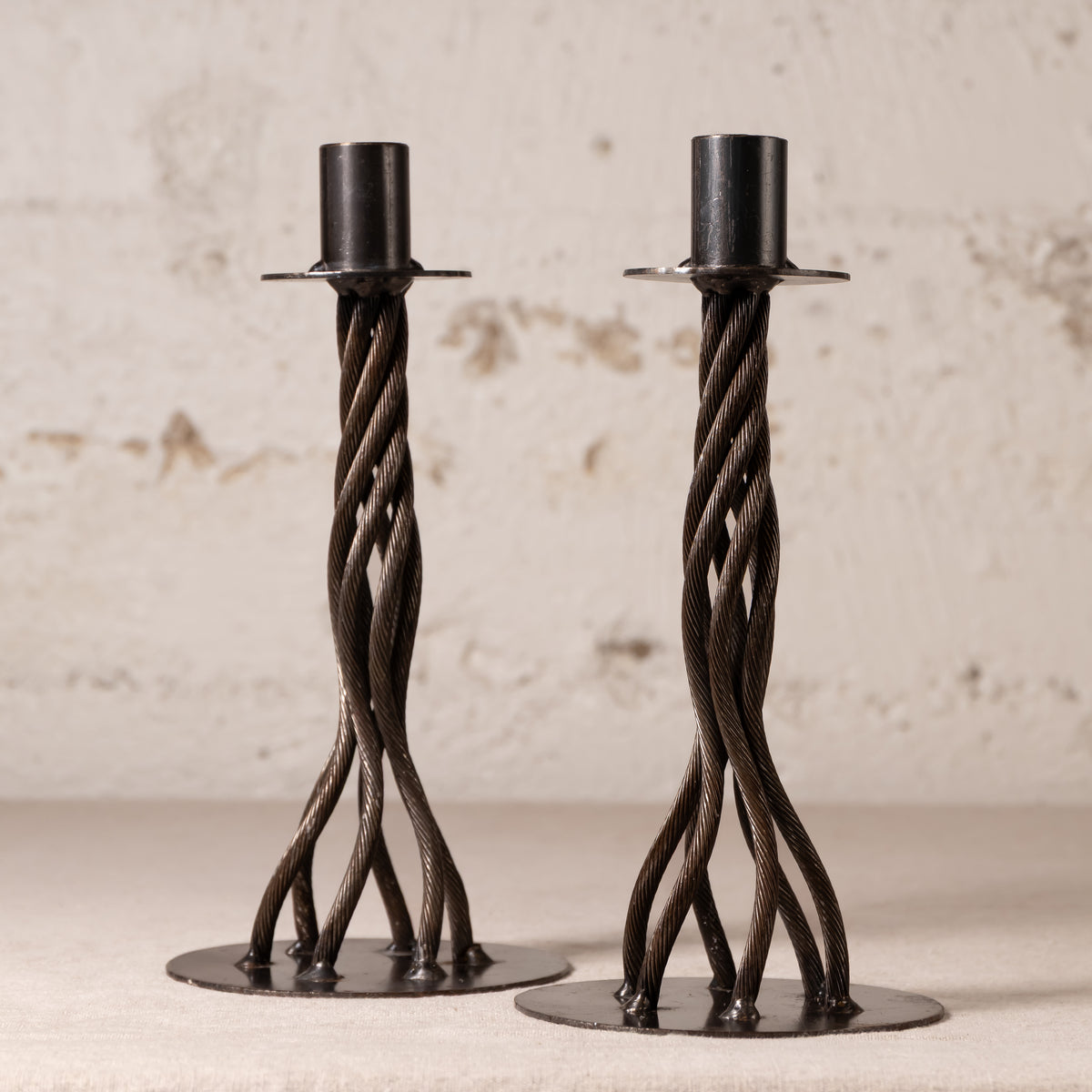 A pair of dark metal twisted candlesticks with circular bases, displayed on a light surface against a textured off-white wall.