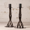 A pair of dark metal twisted candlesticks with circular bases, displayed on a light surface against a textured off-white wall.