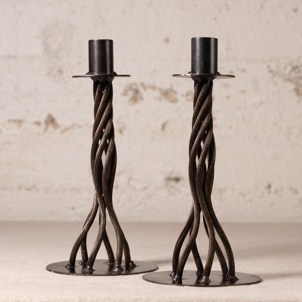A pair of dark metal twisted candlesticks with circular bases, displayed on a light surface against a textured off-white wall.