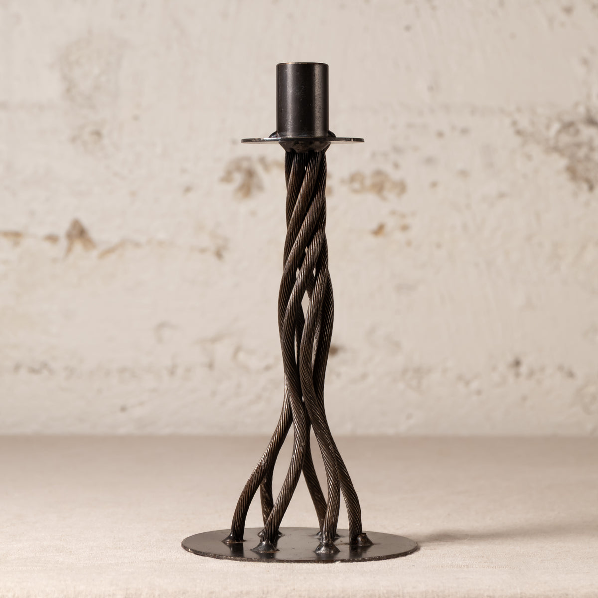 A black metal twisted candlestick holder with a circular base and a cylindrical top, set against a textured off-white wall.
