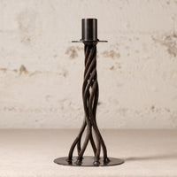 A black metal twisted candlestick holder with a circular base and a cylindrical top, set against a textured off-white wall.