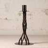 A black metal twisted candlestick holder with a circular base and a cylindrical top, set against a textured off-white wall.