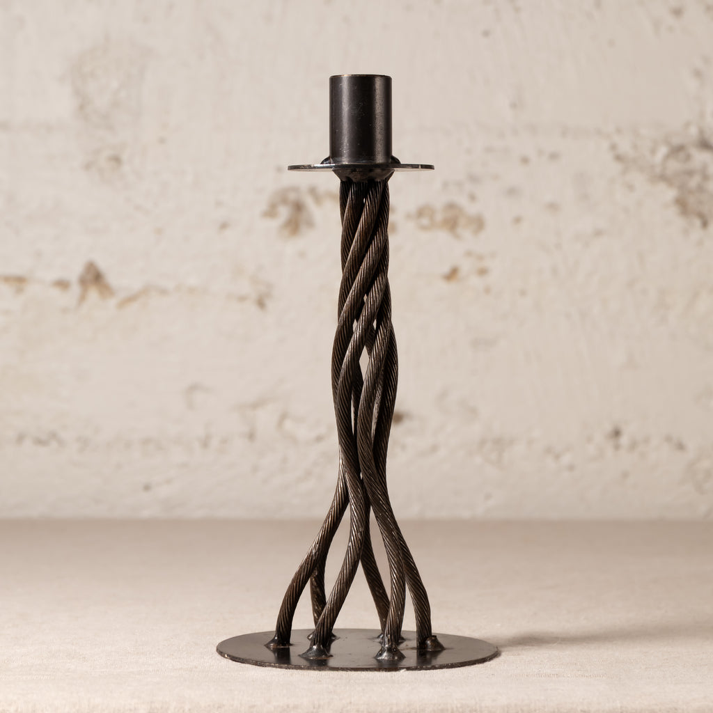 A black metal twisted candlestick holder with a circular base and a cylindrical top, set against a textured off-white wall.