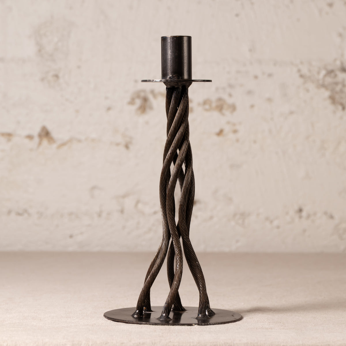 A dark metal twisted candlestick holder with a circular base and a cylindrical top, set against a textured off-white wall.