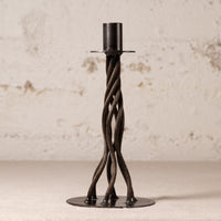 A dark metal twisted candlestick holder with a circular base and a cylindrical top, set against a textured off-white wall.