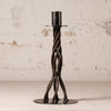 A dark metal twisted candlestick holder with a circular base and a cylindrical top, set against a textured off-white wall.