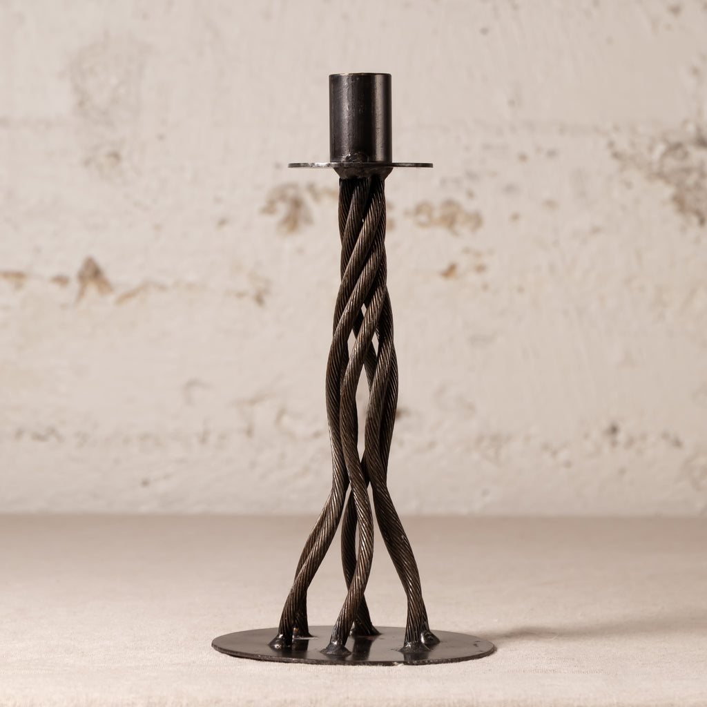 A dark metal twisted candlestick holder with a circular base and a cylindrical top, set against a textured off-white wall.
