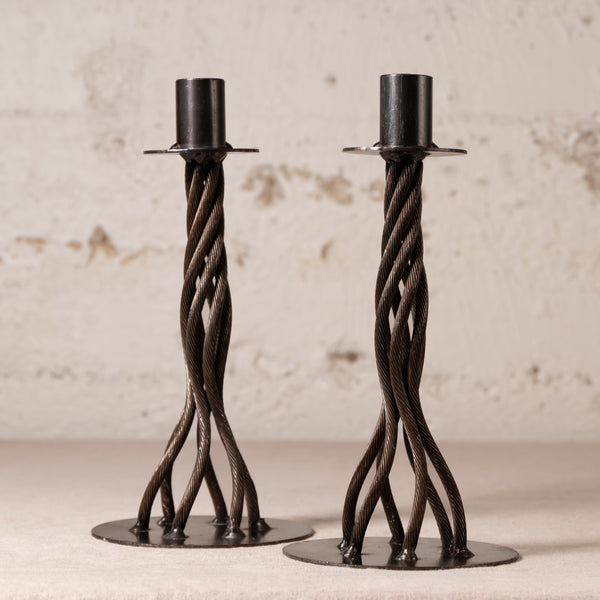 A pair of dark metal twisted candlesticks with circular bases, displayed on a light surface against a textured off-white wall.