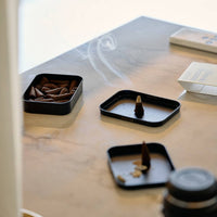 Black square trays with incense cones on a reflective surface