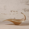 A sculptural, flowing leaf-shaped glass dish in a warm amber-brown tone with a polished metallic gold rim, displayed on a neutral surface against a textured, distressed off-white brick wall.