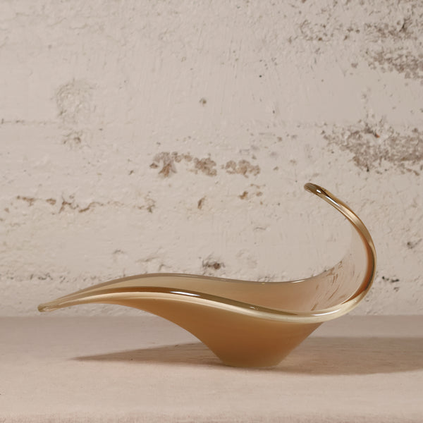 A sculptural, flowing leaf-shaped glass dish in a warm amber-brown tone with a polished metallic gold rim, displayed on a neutral surface against a textured, distressed off-white brick wall.