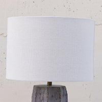 A table lamp with a large white fabric drum shade and a textured gray stone base, shown against a light neutral wall.