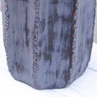 A close-up of a dark charcoal gray textured ceramic pot with vertical ridges and rough, rustic edges, sitting on a white surface.