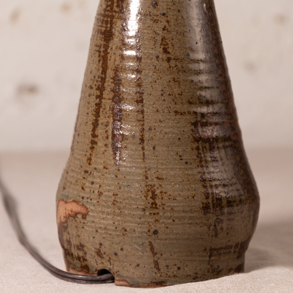 Close-up of a handmade ceramic table lamp with a tapered conical shape, featuring a textured glaze in earthy brown, olive green, and black with a small piece of terracotta showing near the base. A black power cord is visible on the left against a neutral, warm beige background.