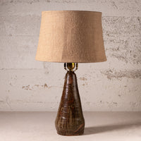 A rustic table lamp with a textured beige burlap drum shade and a dark brown glazed ceramic base, set against a distressed white brick wall.