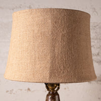 A close-up of a table lamp shade made of natural beige burlap fabric, mounted on a dark brown and gold metal base against a textured off-white wall.