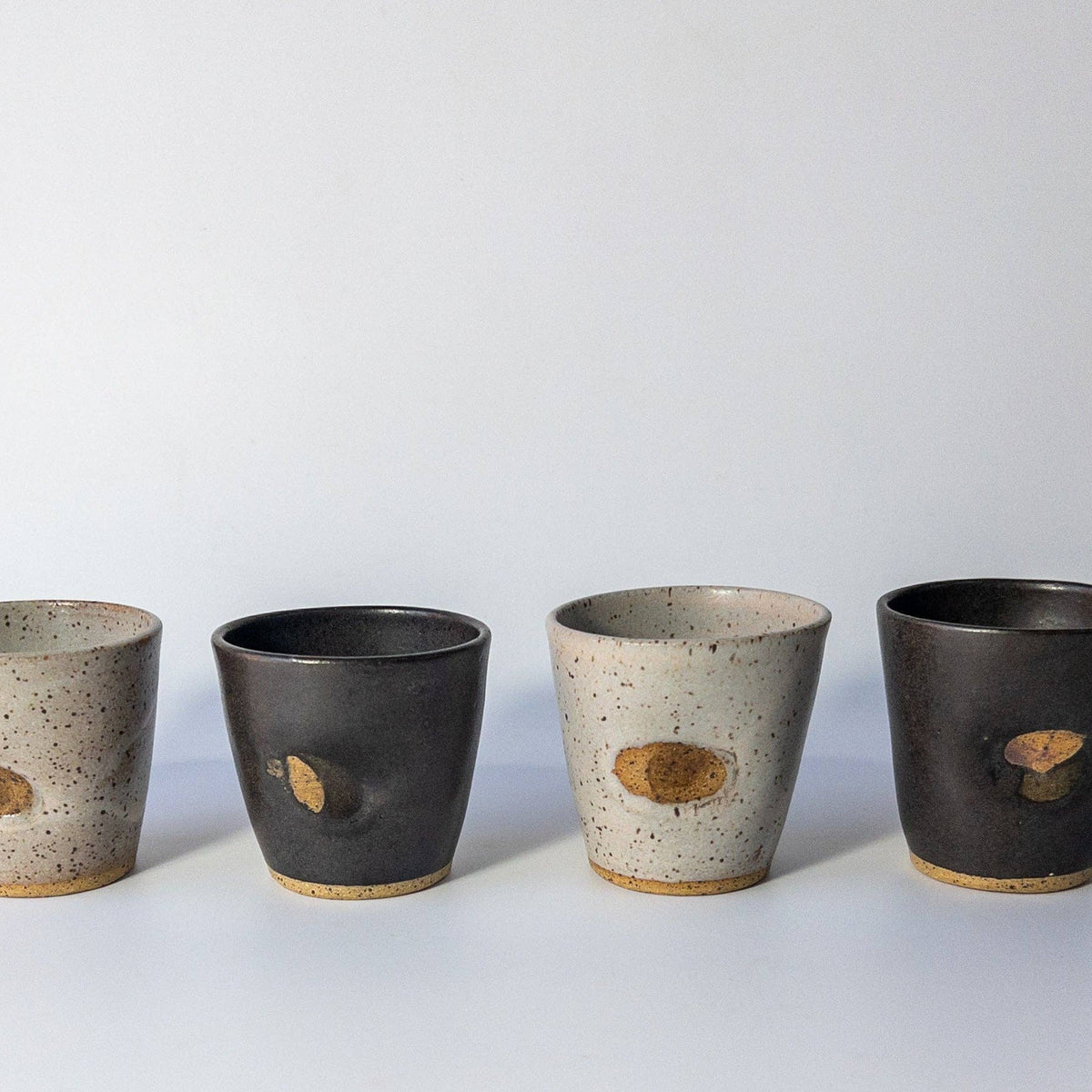 Four handmade ceramic cups lined up on a light gray surface against a white background. Two cups have a speckled light gray and beige glaze with a small oval brown inlay, while the other two have a dark charcoal gray and beige glaze with matching oval inlays.