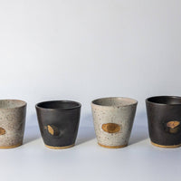 Four handmade ceramic cups lined up on a light gray surface against a white background. Two cups have a speckled light gray and beige glaze with a small oval brown inlay, while the other two have a dark charcoal gray and beige glaze with matching oval inlays.