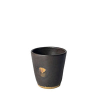 A small dark ceramic cup with a matte black finish, featuring a small gold leaf detail on the front and a natural beige clay base, set against a plain light gray background.