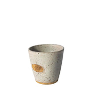 A handmade ceramic cup with a speckled light gray and beige glaze, featuring a small oval brown patch on the side, set against a plain white background.