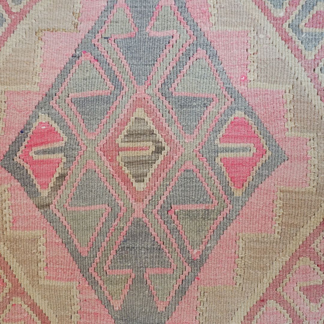 A close-up of a woven textile with a geometric kilim-style pattern in pink, beige, and gray.